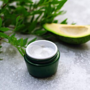 Bowl of body cream with avocado on grey background. Jar of cosmetic cream, leaves and avocado